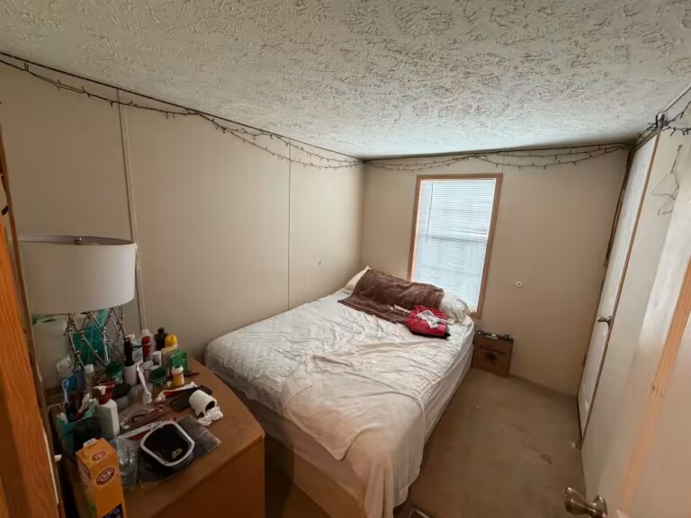 Small bedroom with unmade bed, window with blinds, and cluttered dresser with lamp and toiletries