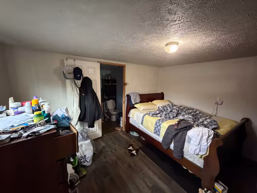 Cluttered bedroom with sleigh bed, messy dresser, coats on door, and bathroom visible through doorway.