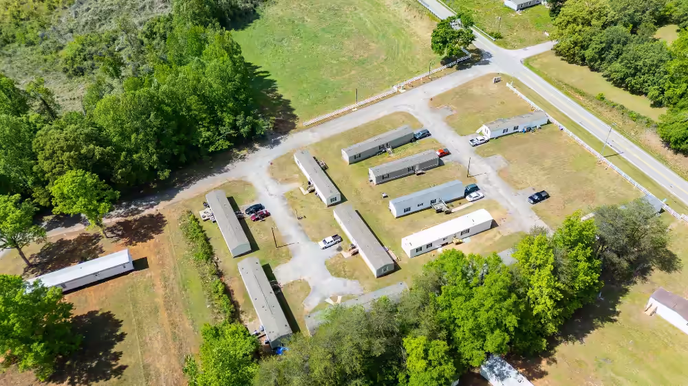 Aerial view of a mobile home park with trailers along gravel roads, surrounded by trees and fields.