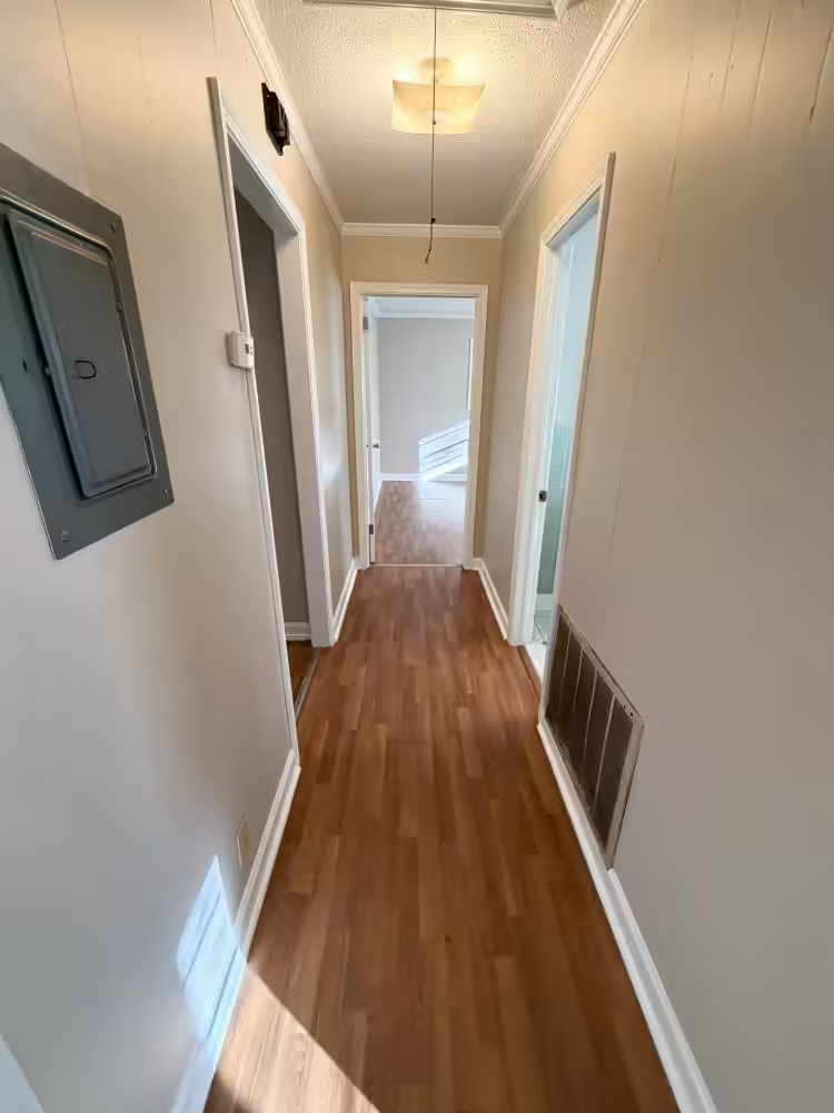 Hallway with wood floors, beige walls, ceiling light, and open doorways leading to rooms.