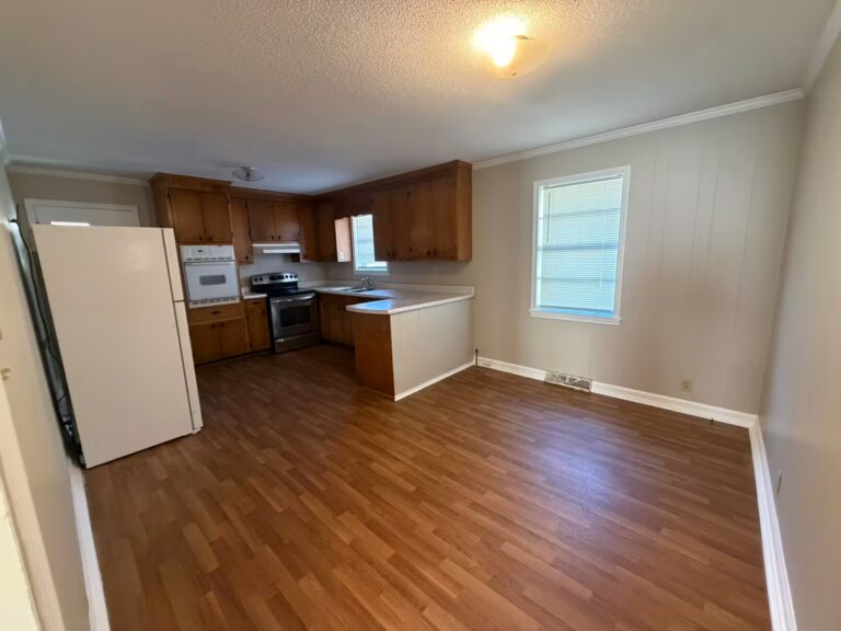 Empty kitchen with wooden cabinets, white refrigerator, stove, and wood-look flooring.